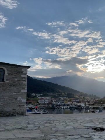 This video presents a serene, panoramic view of a picturesque coastal village, likely Skala Potamias in Greece, during what appears to be late afternoon or early evening. The scene opens with a partial view of a stone building on the left, foregrounding a wide expanse of stone-paved ground.

The midground features a tranquil harbor filled with numerous small boats, including fishing vessels and pleasure crafts, gently bobbing in the calm water. Along the waterfront, several white-washed buildings with terracotta roofs form the heart of the village, nestled at the base of imposing mountains. A few people can be seen casually sitting or walking along the promenade, enjoying the peaceful atmosphere. A yellow vehicle is parked near the waterfront on the right side of the frame towards the end of the video.

The background is dominated by lush, tree-covered mountains that rise dramatically behind the village. The sky is a prominent feature, filled with a mix of fluffy white cumulus clouds and darker, more dramatic stratus clouds. The sun, partially obscured by the clouds, is breaking through, casting striking rays of light across the mountainous landscape and illuminating parts of the village with a soft, golden glow. This creates a beautiful interplay of light and shadow, enhancing the depth and beauty of the scene. The overall impression is one of tranquility and natural beauty, characteristic of a Mediterranean coastal setting.
