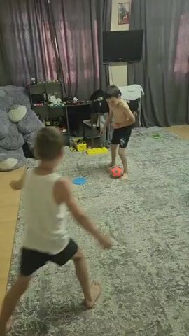 Two young boys are playing on a rug in a room. One boy, wearing a white tank top and black shorts, is facing away from the camera and appears to be jumping or dancing. The other boy, shirtless and wearing black shorts, is standing in the background, looking towards the first boy. There is a large grey teddy bear to the left of the frame, a shelf with various items behind it, and a television mounted on the wall above. The room is lit, suggesting it is daytime, though the weather is not discernible. The overall scene depicts a casual indoor playtime.