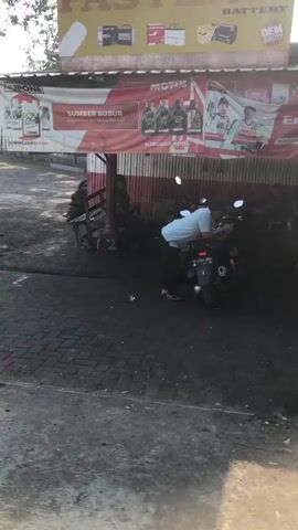 The video shows a street scene with a motorcycle repair shop or service station. In the foreground, a dark, paved area is visible, possibly a driveway or street, with some loose gravel or debris. In the midground, a motorcycle is parked, and a person in a light blue shirt and dark pants is seen near it, appearing to be working on it or preparing to ride it. Another person, wearing dark clothing, is visible behind the motorcycle. To the left, several other individuals are gathered, some leaning against a structure. Behind them, a red and white awning with various advertisements for products like "MOTUL" and "SUMBER SUBUR" is prominent. Above this, a larger sign with the word "FAST" is partially visible, along with images of batteries. The environment appears to be outdoors, with daylight suggesting it's daytime. The overall atmosphere seems to be that of a busy local business.
