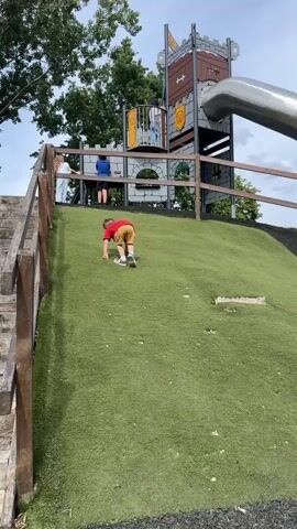 A young child wearing a red shirt and tan shorts is crawling up a grassy hill towards a playground structure designed to look like a castle. The playground has a large metal slide. Several other children and at least one adult are visible on the playground structure at the top of the hill. The environment appears to be an outdoor park with trees and a cloudy sky.