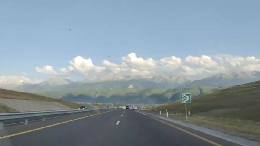 The video shows a view from a car driving on a highway. The car is on a paved road with clear lane markings and guardrails. In the distance, there are mountains covered in green vegetation and snow-capped peaks, with a clear blue sky and scattered white clouds above. The road has other vehicles, including a silver car in front. The surrounding landscape consists of rolling hills and sparse vegetation. There are road signs visible along the roadside.