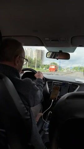 From the backseat of a car, a man with short grey hair and glasses, wearing a dark sweater, is seen driving on a highway. The dashboard shows two phones mounted with chargers plugged in. Outside, buildings and other cars are visible on a cloudy day.