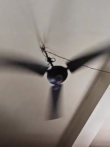 A black ceiling fan with three blades spins rapidly overhead against a cream-colored ceiling. The blades are blurred due to their speed, creating shadows on the ceiling. A thin black wire runs from the fan to the left side of the frame. The fan is suspended from the ceiling by a dark fixture.