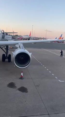 The video shows a close-up view of a white Lufthansa airplane parked on an airport tarmac. The camera pans across the fuselage, highlighting the airline's name in large navy blue lettering and several oval windows. The plane's engine, landing gear, and wing are visible in the foreground. In the background, other airplanes, airport lights, and a clear sky suggest it is either dawn or dusk. A person in a dark uniform is seen standing near the wingtip in the distance. The ground appears to be concrete or asphalt, with some liquid stains visible. A small orange and white traffic cone is placed near the engine. The video appears to be shot from ground level, possibly from the perspective of someone walking towards or boarding the aircraft.