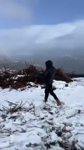 A person in a black hooded jacket, black pants, and tan boots is throwing snowballs near a pile of dead branches. The ground is covered in snow. A rainbow stretches across the sky above a foggy landscape. The person throws a snowball, stumbles slightly, and rights themselves. They resume throwing snowballs.