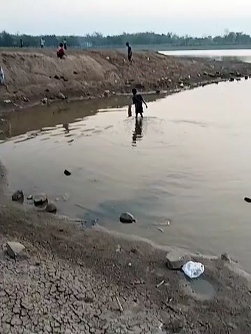 The video shows a group of children playing near a body of water. The water is murky and appears to be shallow. The banks of the water are muddy and cracked, with some rocks and debris scattered along the edge. In the foreground, a child stands in the water, their legs submerged. In the background, other children are visible on a raised dirt bank, some are sitting and others are standing. The sky is overcast and appears to be either dawn or dusk. The overall impression is of a rural or undeveloped area.