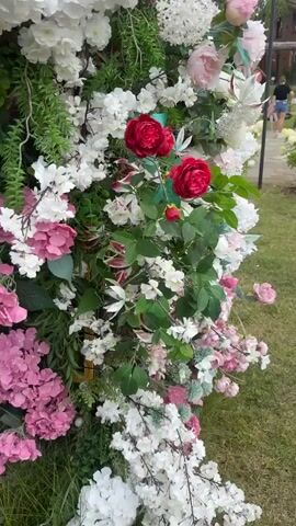 The video shows a close-up view of a dense floral arrangement, likely artificial, with a variety of flowers and greenery. The dominant colors are white, pink, and deep red. Several large red roses are prominently featured in the foreground, with their petals appearing soft and fabric-like. Surrounding them are clusters of smaller white flowers, some resembling cherry blossoms, and larger pink blooms, possibly hydrangeas. Lush green foliage intersperses the flowers, creating a textured and voluminous display. In the blurred background, a grassy area and a hint of a pathway are visible, suggesting an outdoor setting. A person is briefly visible in the far background, walking away from the camera. The lighting appears to be natural daylight, casting soft shadows and highlighting the textures of the flowers.