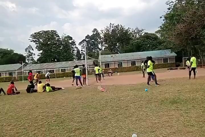 A medium-wide shot shows a volleyball game in progress on a clay court between two teams of young men. They are wearing yellow or red shirts with black shorts. A coach in a dark shirt and blue pants stands on the sidelines, hands on his hips. Several young people sit or lie down on the grassy foreground watching the game. In the background are low, white buildings, trees, and an overcast sky.