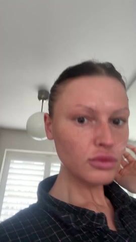 A woman is filming herself in her bedroom. She has her dark hair pulled back into a bun. She's wearing a dark plaid shirt and standing in front of a window with blinds. She says that she loves Sundays because it is a reset day. She moves the camera, showing the room. There is a bed with a brass frame and pink pillows visible. The walls are white and there is a white ceiling.