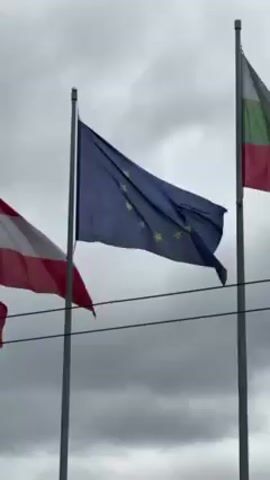 The video shows three flags waving in the wind against a cloudy sky. From left to right, the flags are: the flag of Austria (red-white-red), the flag of the European Union (blue with yellow stars), and the flag of Bulgaria (white-green-red). The flags are attached to silver flagpoles.