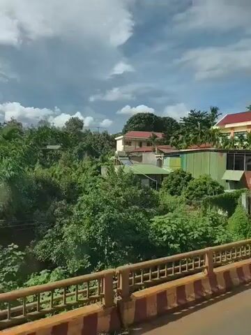 The video shows a view from inside a moving vehicle. The scene outside is a rural or suburban area with buildings, trees, and vegetation. There is a two-story building with a red roof and yellow walls, possibly a school or community center. In front of it, there are smaller buildings, some with green corrugated metal roofs. A motorcycle with a rider is seen on the road. There are also fences, a bridge railing, and a street sign. The sky is partly cloudy.