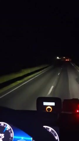 The video captures a first-person perspective from the driver's seat of a vehicle traveling on a dark road at night in Punghina, Romania. The road extends straight ahead into the distance, with the vehicle's headlights illuminating the immediate path. On either side of the road, dark, unlit fields are visible.

Periodically throughout the video, bright flashes of lightning illuminate the dark sky and the surrounding landscape, revealing a wide-open area with no immediate structures. The lightning briefly turns the night sky into a bright, cloudy expanse.

Inside the vehicle, parts of the dashboard are visible in the foreground. On the left, a portion of a speedometer or tachometer dial with blue and white markings is seen. In the center of the dashboard, an electronic device with a small screen and an orange circular light or button is mounted. To its right, another electronic device with two red lights is visible. The taillights of other vehicles can be seen in the distance ahead on the road. No legible text is visible on any of the devices or elsewhere in the video.
