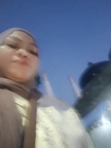 The video shows a young girl wearing a hijab and a light-colored outfit. She is looking at the camera and adjusting her hijab. In the background, there is a tall white structure that appears to be a minaret, suggesting the presence of a mosque. There are also trees, parked motorcycles, and other people in the distance. The sky is a deep blue, indicating it is either dusk or dawn. The video is slightly blurry and shaky, giving the impression that it was filmed while someone was moving.