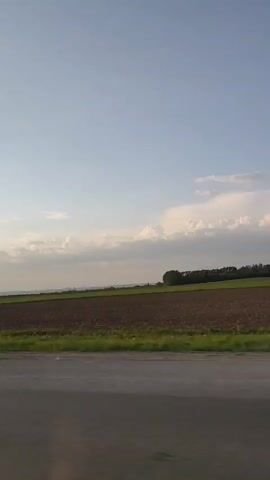 The video shows a landscape view from the passenger seat of a car driving on a road. The scenery includes a cultivated field, a line of trees, and a partly cloudy sky. The road stretches out in the direction the car is traveling. In the reflection in the side mirror you can see the setting sun. Loud music is playing.