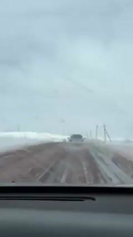 The video shows a view from inside a car driving on a snow-covered road. Another car is visible ahead, driving in the same direction. The road appears to be a reddish-brown color beneath the snow and ice. Power lines are visible on the side of the road. The sky is overcast and gray, suggesting a winter day. The focus is on the road conditions and the car ahead.