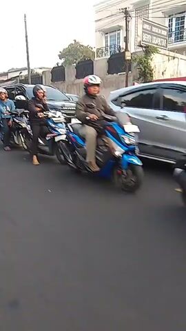 The video shows a street scene with multiple vehicles and people. In the foreground, several motorcycles are traveling on a road. Some are occupied by one or two individuals wearing helmets. In the background, there are cars, including a dark SUV and a white car, as well as a large white truck. Buildings and trees are visible along the side of the road. The scene appears to be a busy street with moderate traffic. The camera is moving along with the motorcycles, giving a sense of motion.