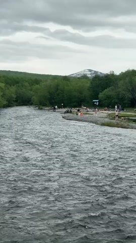 This is a wide shot of a wide, fast-flowing river with choppy water under a cloudy sky. On the far bank, there is a gravelly shore with scattered trees and bushes. Several people are visible on the shore, some sitting, some standing, and some appearing to be in the water. Some people are wearing swimsuits. In the background, there is a dense line of green trees, and beyond that, a distant mountain peak with some snow on its summit. The overall impression is of a natural, outdoor setting with people enjoying the water.