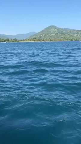 The video shows an expanse of blue water with small waves. In the distance, a coastline is visible, dotted with trees and a few structures. Behind the coastline, a green, hilly landscape rises towards a clear, blue sky. A person speaks in the background, but they are not visible.
