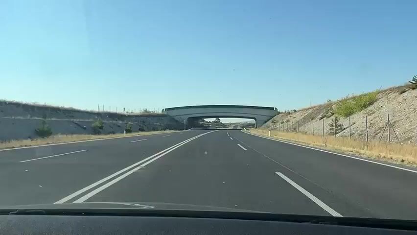 The video shows a first-person perspective of driving on a highway under a clear blue sky. The road is paved and has white lane markings. On either side of the road, there are gently sloping hills covered with dry grass and some scattered bushes and small trees. A large concrete overpass with a green railing runs across the road ahead. The overpass is supported by substantial concrete pillars and appears to be designed to allow passage of animals or vehicles underneath. The camera is positioned inside a vehicle, likely from the driver's viewpoint, showing the dashboard in the foreground. As the vehicle approaches the overpass, the view transitions to driving underneath it. Beyond the overpass, the highway continues straight, with more hills visible in the distance. There are no other vehicles or people clearly visible in the frame.