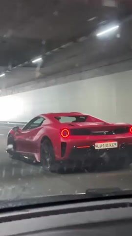 The video shows a red Ferrari driving through a tunnel and then exiting onto a road. The car has black wheels and a license plate that reads "LU-131932." The tunnel has concrete walls and is lit by fluorescent lights. The road outside the tunnel is lined with trees and has a black fence on the right side. The video is shot from inside another car.