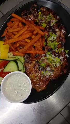 A close-up shot from above shows a black plate of food on a stainless steel counter. The plate contains a pile of crispy, orange sweet potato fries, a small salad with lettuce, cucumber slices, cherry tomatoes, and a wedge of yellow cheese, and a small white ramekin filled with white dipping sauce. The largest portion of the plate is occupied by grilled meat, likely ribs or chicken, glazed with a dark, glossy sauce and sprinkled with chopped green herbs and white onions. The lighting is somewhat dim, and the background is blurred, suggesting a restaurant kitchen environment. There are no people visible in the frame.