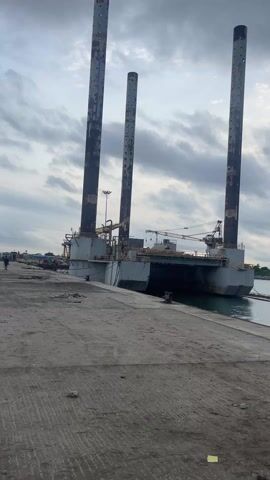 A static shot shows a large, gray, barge-like vessel docked at a concrete pier under a cloudy sky. The vessel has three tall, cylindrical legs extending upwards, showing signs of rust and wear. On the deck of the vessel, various structures, pipes, and a crane are visible. A person walks on the pier to the left of the vessel. The water is calm and reflects the cloudy sky. The overall impression is of an industrial or maritime setting.