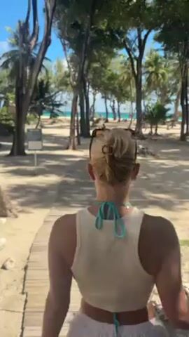 A light skinned woman with a blonde bun walks toward the beach on a wooden path, framed by trees and sand. She turns, waves, and continues walking. She is wearing white shorts and a light yellow tank top with a blue bikini string. She carries a small purse. The beach and turquoise water are visible in the background.
