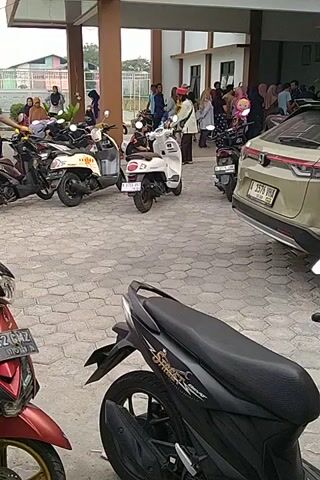The video shows a parking lot with several motorcycles and a car. A group of people are standing near the car, some of them appear to be women wearing headscarves. One person, a man, is walking towards the motorcycles. The ground is paved with interlocking stones. The scene appears to be outdoors, under a covered area with support beams. The lighting suggests it is daytime.