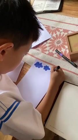 The video shows a close-up view of two children and an adult sitting at a table, engaged in drawing and reading. The scene is brightly lit, suggesting an indoor setting, possibly a home or classroom. In the foreground, a young boy with short dark hair is wearing a white shirt with blue stripes. He is holding a black marker and drawing blue shapes, which appear to be clouds, on a white sheet of paper. To his right, a box of colorful markers is visible. Further back, another child, wearing a light-colored t-shirt, is also drawing on a piece of paper, holding a black marker. An adult, wearing a plaid shirt and glasses, is seated behind the children, engrossed in reading a book. Several open books and notebooks are scattered on the table, along with more markers. The table is covered with a patterned cloth. The overall atmosphere appears to be one of focused activity and shared learning.