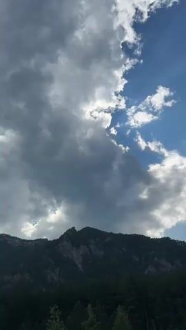 This is a vertical, full shot video of a mountainous landscape in Ust'-Muny, Russia. The scene is dominated by a dramatic sky with a mix of dark, heavy clouds on the left and bright blue sky with scattered white clouds on the right. Sunbeams are visible piercing through the clouds on the right side, indicating a change in weather or a break in the clouds.

The lower half of the frame features a dark, densely forested mountain range. The trees are predominantly evergreen, creating a dark green mass against the lighter sky. The mountain itself has a rugged, rocky peak visible above the treeline on the left side.

The time of day appears to be late afternoon or early evening, given the angle of the light and the contrast between the dark clouds and the illuminated sky. The weather seems dynamic, possibly transitioning from rain or a storm to clearer skies. The emotion conveyed is one of natural grandeur and the power of the elements. There is no visible text in the media.