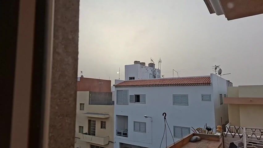 The video shows a view of rooftops of buildings in an urban setting under an overcast sky. The sun appears as a hazy white disc in the sky, barely visible through the cloud cover. The buildings are mostly white and beige, with some having red tile roofs. There are also antennas visible on top of some of the buildings. The video seems to have been taken from an elevated position, possibly from a window or a higher floor within a building. The foreground of the frame includes part of a wall, suggesting the point of view is from inside a building. There are no people visible in the video.