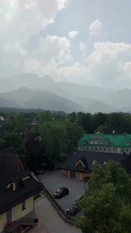 The video pans over a scenic view of a town nestled at the foothills of misty mountains. In the foreground, there are several buildings with dark, sloped roofs, some featuring dormer windows. One prominent yellow building with a dark roof is on the left. A paved road with parked cars, including a black sedan and a dark-colored SUV, winds through the scene. Beyond the buildings, a dense canopy of green trees fills the landscape, interspersed with glimpses of more houses. The background is dominated by the majestic, hazy outlines of several mountain ranges, fading into the distance. The sky above is partly cloudy, with a mix of bright sunlight and soft shadows.