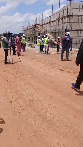 The video shows a construction site with scaffolding surrounding a building. Several people are present, most wearing safety vests and hard hats. One person in the foreground is wearing a black garment and a yellow safety vest, walking away from the camera. In the background, a group of people are gathered, some appearing to be observing the construction. Another person is seen taking a photo with a phone. The ground is dirt and gravel, and the sky is cloudy with patches of blue. Another person in a blue hard hat and yellow vest walks by the camera in the middle ground. The scene suggests an inspection or a tour of the construction site.