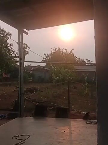 THE VIDEO SHOWS A SUNRISE OR SUNSET OVER A RURAL LANDSCAPE. THE CAMERA IS POSITIONED UNDER A ROOFED STRUCTURE, LOOKING OUTWARD. IN THE FOREGROUND, THERE IS A CONCRETE FLOOR WITH SOME OBJECTS SCATTERED ON IT, INCLUDING WHAT APPEARS TO BE A HOSE AND SOME PIECES OF METAL OR WOOD. THE MIDDLE GROUND FEATURES A YARD WITH TREES, BUSHES, AND SMALL BUILDINGS. THE BACKGROUND IS DOMINATED BY THE SKY AND THE SUN, WHICH IS LOW ON THE HORIZON AND CASTING A WARM, GOLDEN LIGHT. THE OVERALL SCENE IS PEACEFUL AND SERENE.