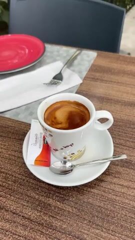 The video shows a close-up shot of an espresso cup and saucer on a wooden table. A packet of sugar and a spoon are placed on the saucer. In the background, a red plate and a fork are visible on a placemat. Beyond the table, there is a railing, chairs, and lush green trees and foliage. The scene appears to be an outdoor seating area, possibly at a cafe or restaurant with a view of nature. The lighting suggests it is daytime. No people are visible in the frame.