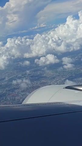 This is a first-person perspective video taken from an airplane window. The camera is angled downwards, showing a view of clouds and a sprawling city below. The wing of the airplane and part of its engine are visible in the foreground, taking up the bottom portion of the frame. The city appears densely populated with buildings and infrastructure, with some signs of green spaces. The sky is partly cloudy with large, fluffy white cumulus clouds interspersed with blue sky. The overall lighting suggests it is daytime, possibly late afternoon given the angle of the light. The video appears to be a short clip, showing the view from the aircraft as it cruises at altitude.