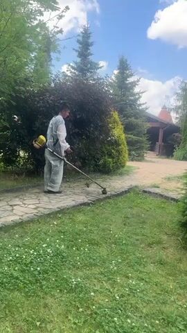 A person in a white hazmat suit is operating a string trimmer on a paved walkway. The person is facing away from the camera and appears to be trimming the grass along the edge of the walkway. The setting is outdoors with trees, bushes, and a building visible in the background. The sky is blue with some clouds. The video is short and appears to be a single, continuous shot.