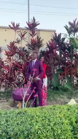 A man and woman are posing for photographs in an outdoor setting. The man is wearing a suit, and the woman is wearing a red and floral outfit. They are standing close together, partially obscured by red leafy plants. A photographer stands in the foreground, taking pictures of the couple with a camera. A green hedge separates the photographer from the couple. A security camera is mounted on a pole in the background, and a residential building can be seen. The sky is blue.