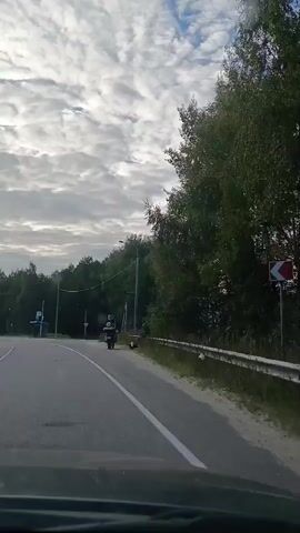 The video shows a view from inside a vehicle driving on a paved road. The road has a white dashed line on the right side and a solid white line on the left. On the left side of the road, there are trees and a blue bus stop with a sign. On the right side of the road, there is a metal fence and more trees. The sky is cloudy. A motorcycle with a person wearing a helmet is parked on the side of the road with a person standing next to it. Another person is seen sitting near the motorcycle. Further down the road, a car passes by. The overall scene appears to be a rural or suburban setting.