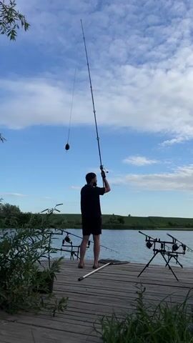 A person wearing a black t-shirt and black shorts stands on a wooden dock, facing away from the camera, holding a fishing rod and casting it into a lake under a partly cloudy sky. There are fishing rods in rod holders on the dock, and fishing nets are visible. Lush green foliage is in the foreground and background.