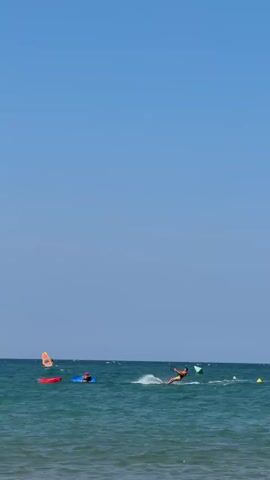 The video captures a bright, sunny day at sea. The sky is a clear, unbroken blue. The ocean is a vibrant teal, with gentle waves rolling towards the shore. In the distance, the horizon line is sharp and distinct.

The main focus of the video is a person kitesurfing. They are wearing yellow shorts and are actively engaged in the sport, with a spray of water visible behind them as they move across the water. The kite they are using is not clearly visible.

Scattered around the water are several other objects. To the left of the kitesurfer, there is a windsurf sail, orange and triangular in shape, attached to a board. Further to the left, a small dark object, possibly another kite, is visible in the distance. In the foreground, closer to the left edge of the frame, there are two objects floating on the water: one is a red rectangular inflatable, and the other is a dark blue inflatable. There are also some small, distant buoys.

The overall impression is one of an active and sunny day at the beach, with various water sports activities taking place.