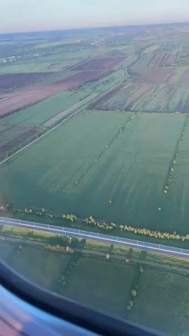 The video shows a view from an airplane window. Below, the landscape is divided into rectangular fields, some green and some brown. A highway with a few vehicles runs horizontally across the frame. The long shadows of trees stretch across the green fields. A smaller road intersects the highway.