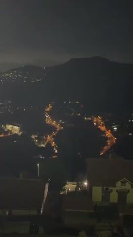 A nighttime aerial view shows a town nestled in a valley below a mountain range. The town's lights create winding lines of illumination that follow the contours of the landscape. In the foreground, the rooftops of several buildings are visible, their tile work dark against the night sky. The overall impression is one of a tranquil, albeit dimly lit, urban spread against a natural backdrop.