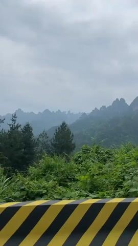 The video shows a view of mountainous terrain under an overcast sky, captured from a moving vehicle. The mountains are covered in lush green vegetation, and their peaks are jagged and distinct. In the foreground, there's a safety barrier with black and yellow diagonal stripes. As the camera moves, a metal structure resembling a watchtower or platform is visible on the left side. Utility poles with electrical wires also appear intermittently in the shot. The overall impression is a scenic landscape observed during travel.