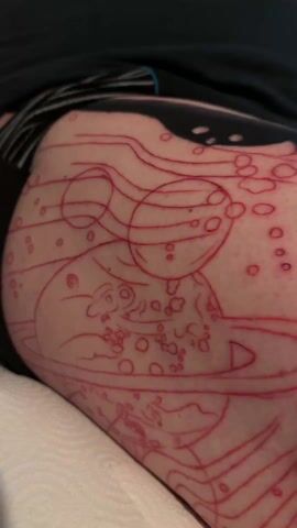 The video shows a person's leg with a tattoo being applied. The leg has a faint red outline of a design, which appears to include planets and other circular shapes. A tattoo artist has already partially completed a tattoo on the leg featuring a face with red and white stripes, with black stripes emanating from the face. The leg rests on a white cloth surface. The person wearing the shorts is not visible. The focus remains on the leg and tattoo.