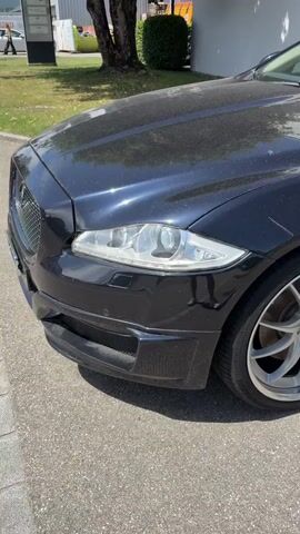 The video shows a dark-colored Jaguar XJ with the model designation "STARTECH Automatic". The car is parked outdoors on asphalt, with some grass and trees visible in the background. The camera pans across the front and side of the vehicle, focusing on the headlights, wheels, and a sign displayed on the windshield. The sign provides details about the car, including its model, year, mileage (68,000 km), color, transmission type (automatic sequential), fuel type (Diesel), and price (CHF 49,500). The car appears to be for sale as a showroom car. The exterior of the car is dusty.