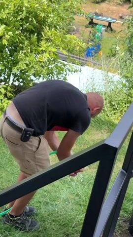 A bald man wearing a black t-shirt and khaki shorts is outside watering plants with a green hose. He is bent over, holding the hose with both hands and aiming it downwards. He is standing on a grassy slope next to a black railing. In the background, there are trees and a pool.