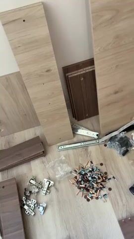 The video shows the assembly process of a large brown wardrobe. The scene initially presents the various components of the wardrobe scattered on a light wooden floor, including wooden panels, hinges, screws, and a power drill. It then pans upwards to reveal the completed wardrobe standing against a light-colored wall. The wardrobe consists of four doors with silver handles and a wood-grain finish.