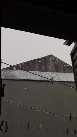 The video captures a view from an indoor space, likely a window or doorway, looking out onto an exterior scene in Monte Grande, Argentina. The primary subject is a grey building with a distinct triangular roof, partially obscured by what appears to be another grey wall or structure in the foreground. Several clotheslines with various colored clothespins (brown, green, black) are strung across the lower and middle sections of the frame. It is raining, with individual raindrops visibly falling and accumulating on surfaces, including the clotheslines. The sky is overcast and grey, indicating cloudy weather. The overall impression is that of a quiet, rainy day. No people are visible, and there is no discernible text.