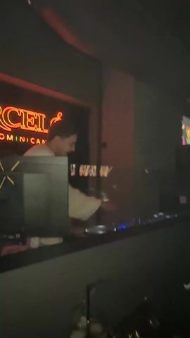 The video shows a DJ playing music in a nightclub with a neon sign in the background. The DJ is a young man wearing a white sweater and operating a Pioneer DJ controller. The nightclub is dimly lit with blue and red lights. The crowd is diverse and enjoying the music and dancing. Some people are seen mingling and chatting. The camera moves around the room showing different aspects of the scene, sometimes focusing on individuals in the crowd. The atmosphere is lively and energetic.
